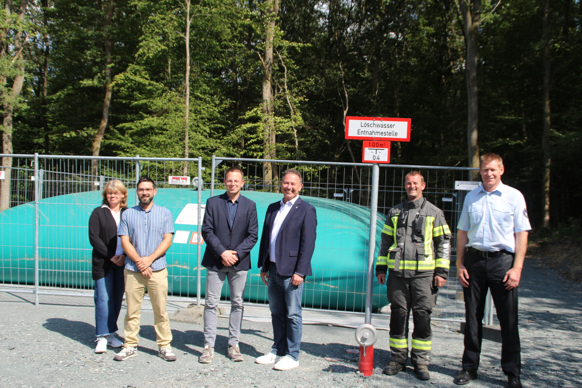 Im Hintergrund ist ein großes Löschwasserkissen im Wald. Im Vordergrund sind die beiden Bürgermeister und die Verantwortlichen aus Verwaltung und der Feuerwehr zu sehen.