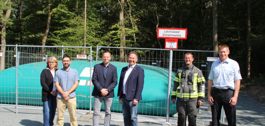 Im Hintergrund ist ein großes Löschwasserkissen im Wald. Im Vordergrund sind die beiden Bürgermeister und die Verantwortlichen aus Verwaltung und der Feuerwehr zu sehen.