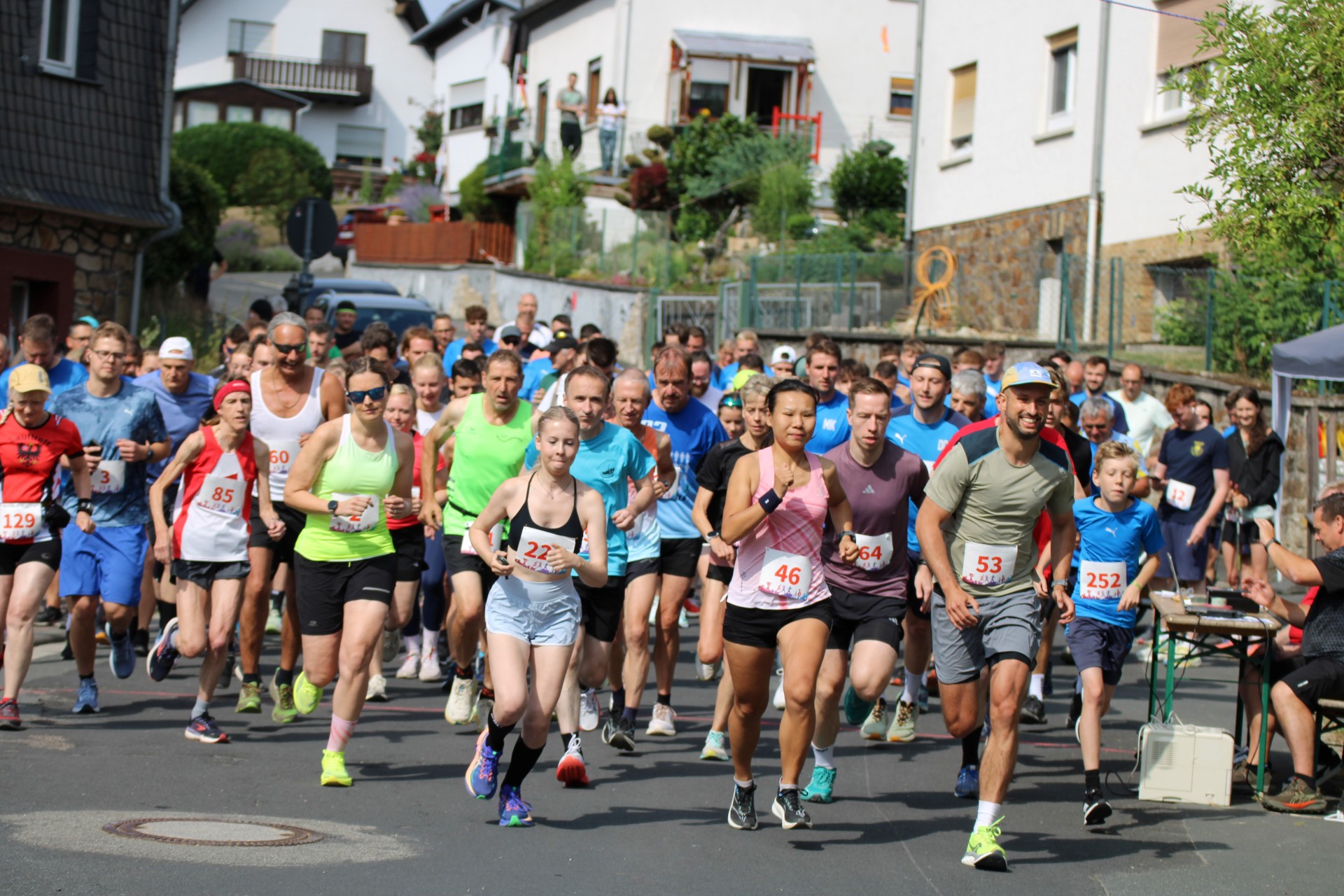 Teilnehmerinnen und Teilnehmer starten gemeinsam bei einem Volkslauf in einer Ortschaft und laufen motiviert auf der Straße los.